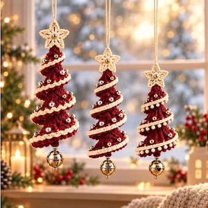 Op de afbeelding: Drie hangende kerstboomornamenten gemaakt van donkerrood haakgaren. Elke boom is voorzien van een spiraalvormige crèmekleurige gehaakte rand, kleine zilveren kralen, een crèmekleurige gehaakte ster aan de top en een gouden belletje aan de onderkant.
