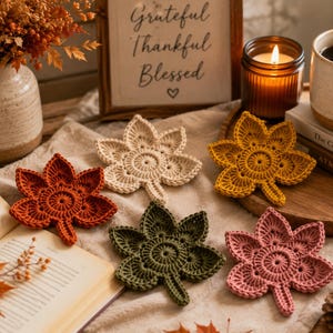 Peut inclure: Cinq dessous de verre crochetés en forme de feuille, de couleur crème, jaune moutarde, orange brûlé, vert olive et vieux rose. Chaque dessous de verre présente un centre circulaire avec des pointes rayonnantes. Ils sont posés sur un tissu clair.