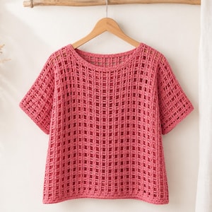 May include: A short-sleeved, round-neck top crafted from a vibrant pink yarn in an open, grid-like crochet pattern. The garment features a boxy silhouette with a straight hemline and is displayed on a wooden hanger, showcasing the intricate texture.