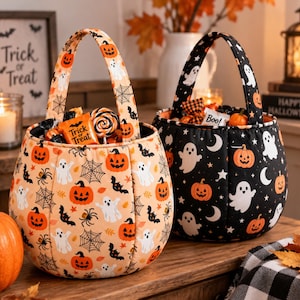 May include: Two fabric Halloween candy buckets with handles. One bucket is light orange with a pattern of pumpkins, ghosts, spiders, and webs. The other bucket is black with a pattern of white ghosts, orange pumpkins, stars, and crescent moons.