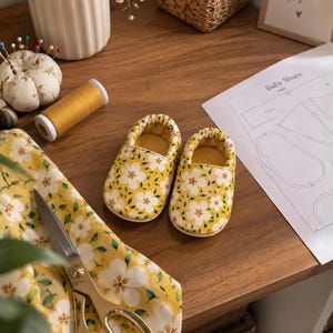 May include: A pair of baby shoes featuring a yellow fabric with a white floral pattern and green leaves. The shoes have a rounded toe and an elasticized opening. Beside the shoes are matching fabric, gold scissors, yellow thread, and a paper pattern.