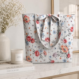 May include: A white quilted tote bag featuring a floral pattern with red, orange, and blue flowers and green leaves. The bag has two vertical fabric straps that tie into a bow at the top center, with vertical stitching throughout the exterior.