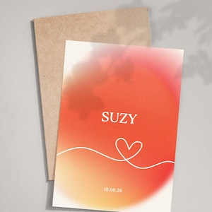 Op de afbeelding: Een wenskaart met een oranje en witte gradiënt. Het woord "SUZY" staat bovenaan, met een wit hart en een datum eronder. Achter de kaart zit een bruine envelop.