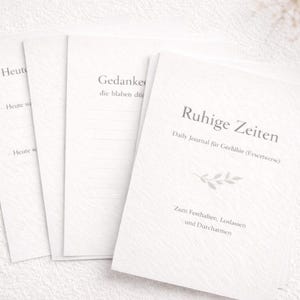 Op de afbeelding: Een stapel witte tijdschriften met Duitse tekst, waaronder "Ruhige Zeiten" en "Daily Journal". De tijdschriften hebben een getextureerde omslag en liggen op een wit oppervlak.