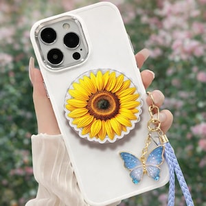 May include: A white smartphone case featuring a large, vibrant yellow sunflower graphic centred on the back. Attached to the side is a gold-toned metal charm shaped like a blue butterfly with glittery accents, connected to a braided light blue strap.