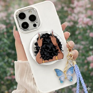 May include: White phone case with a pop-up grip featuring a cartoon illustration of a person with curly black hair and raised fingers. A blue butterfly charm hangs from the case with a light blue lanyard.