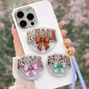 May include: A white phone case with three "MAMA" phone grips. The grips feature a leopard print design with bows in brown, pink, and teal. A blue wrist strap is attached to the phone case.