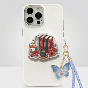 May include: A white smartphone case featuring a sticker of four books labeled Twisted Fairy Tales, Dark Romance, Trigger Warnings, and Smutty Vibes, alongside a lit candle, a mug, and red roses, with a blue butterfly charm on a braided strap.