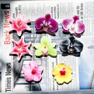 May include: A collection of colorful flower-shaped hair clips. The clips feature various designs, including pink lilies, purple orchids, and yellow hibiscus. The background is a newspaper.