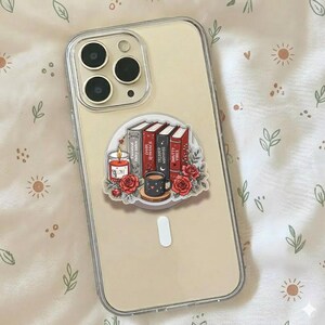 May include: A clear smartphone case featuring a sticker with a stack of four books titled Twisted Fairy Tales, Dark Romance, Trigger Warnings, and Smutty Vibes, accompanied by a lit candle, a mug, and red roses with green leaves.
