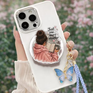 May include: White phone case with a graphic of a person reading a book and the text "Just one more chapter." A blue butterfly charm hangs from the case. The phone case has a clear camera lens protector.