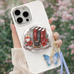 May include: A white smartphone case featuring a circular graphic of four books titled Twisted Fairy Tales, Dark Romance, Trigger Warnings, and Smutty Vibes, alongside a lit candle, a coffee mug, and red roses, with a blue butterfly charm attached.
