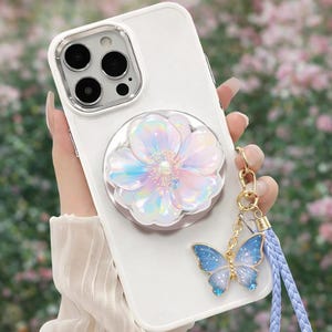 以下が含まれることがあります： 白い携帯電話ケース。虹色の花の形をした電話グリップが付いています。青い蝶のゴールドチャームと水色のリストストラップが付属しています。携帯電話ケースには透明なカメラレンズプロテクターが付いています。