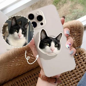 May include: A light gray phone case featuring a black and white cat face design. The case has a built-in pop-up grip with the same cat design. The phone case is held by a person.