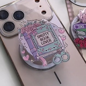 May include: A clear phone grip featuring a book design with the text "Smutty Book Lover." The book is light blue with a purple spine, pink ribbon, and handcuffs. Pink roses and hearts surround the book.