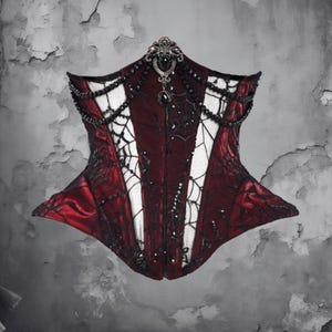 May include: A burgundy corset with black spiderweb and beaded accents. The bodice features a decorative jeweled clasp at the top and a central panel of white fabric. The corset is a gothic-inspired garment.