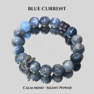 Puede incluir: Una pulsera de cuentas azules y grises con un dije de serpiente plateada. Las palabras "BLUE CURRENT" y "CALM MIND • SILENT POWER" están impresas encima y debajo de la pulsera.