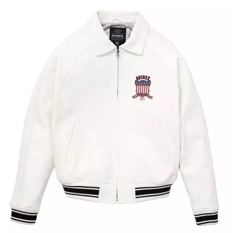 May include: White jacket with a collar and a zipper closure. The jacket features a black and white striped hem and cuffs. An emblem with red, white, and blue details is on the chest. The brand name "AVIREX" is visible.