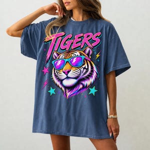 May include: A denim blue oversized t-shirt featuring a graphic print of a tiger wearing bright blue and pink sunglasses. The word TIGERS is printed in a bold pink font above the tiger, surrounded by colorful lightning bolts and star shapes.