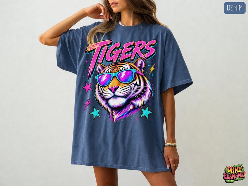 May include: A denim blue oversized t-shirt featuring a graphic print of a tiger wearing bright blue and pink sunglasses. The word TIGERS is printed in a bold pink font above the tiger, surrounded by colorful lightning bolts and star shapes.