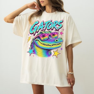 May include: An ivory t-shirt featuring a graphic of a rainbow-colored alligator wearing sunglasses with palm tree reflections. The text GATORS is printed in a bold blue font above the alligator, surrounded by colorful stars and lightning bolts.