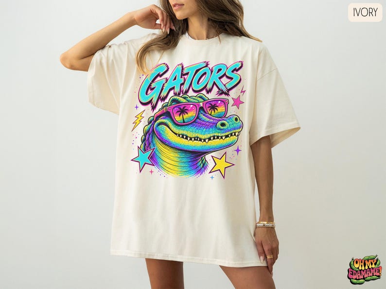 May include: An ivory t-shirt featuring a graphic of a rainbow-colored alligator wearing sunglasses with palm tree reflections. The text GATORS is printed in a bold blue font above the alligator, surrounded by colorful stars and lightning bolts.
