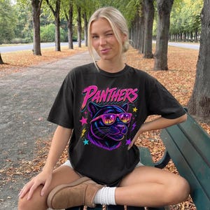 May include: A black short-sleeve t-shirt featuring a graphic of a panther wearing pink and orange sunglasses with palm tree silhouettes. The word PANTHERS is printed in a stylized pink font above the panther, surrounded by colorful stars and lightning bolts.