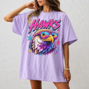 May include: An orchid-colored t-shirt featuring a graphic of an eagle wearing sunglasses with palm tree reflections. The word HAWKS is printed in a stylized pink and blue font above the eagle, surrounded by colorful stars and lightning bolt accents.