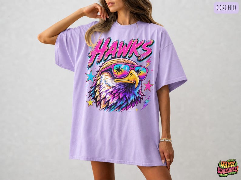 May include: An orchid-colored t-shirt featuring a graphic of an eagle wearing sunglasses with palm tree reflections. The word HAWKS is printed in a stylized pink and blue font above the eagle, surrounded by colorful stars and lightning bolt accents.