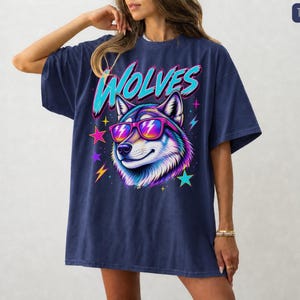 May include: A true navy short-sleeve t-shirt featuring a graphic of a wolf wearing pink and orange sunglasses. The word WOLVES is printed in large cyan letters above the wolf. The design includes colorful stars and lightning bolt accents.