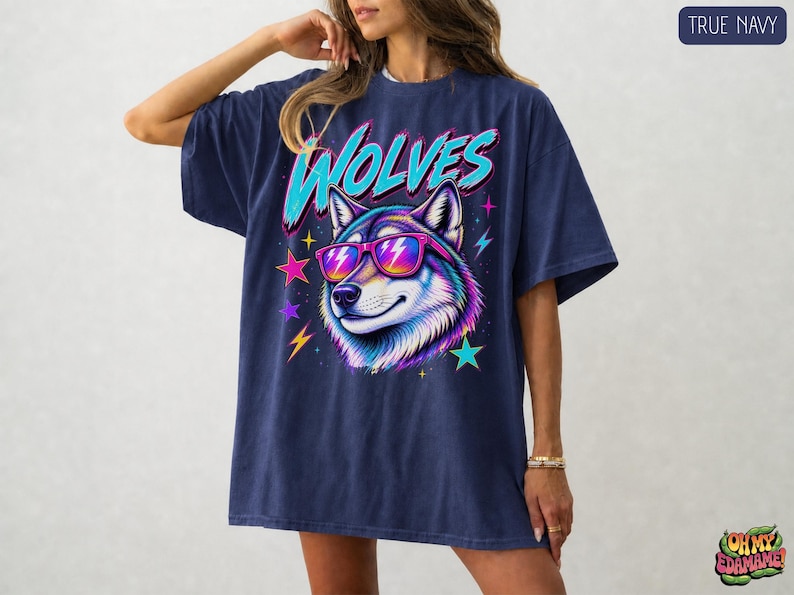 May include: A true navy short-sleeve t-shirt featuring a graphic of a wolf wearing pink and orange sunglasses. The word WOLVES is printed in large cyan letters above the wolf. The design includes colorful stars and lightning bolt accents.