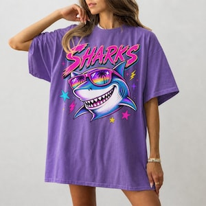 May include: A violet oversized t-shirt featuring a graphic print of a shark wearing rainbow-tinted sunglasses with palm tree reflections. The word SHARKS is printed in a stylized pink font above the shark, surrounded by colorful stars and lightning bolts.