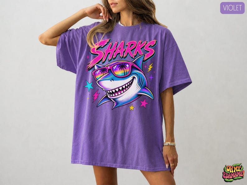 May include: A violet oversized t-shirt featuring a graphic print of a shark wearing rainbow-tinted sunglasses with palm tree reflections. The word SHARKS is printed in a stylized pink font above the shark, surrounded by colorful stars and lightning bolts.