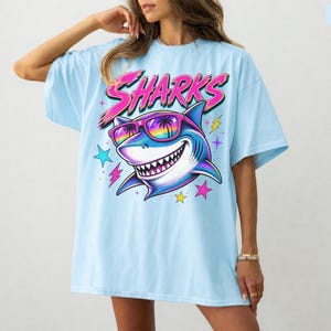 May include: A light blue t-shirt featuring a graphic of a shark wearing rainbow-tinted sunglasses with palm tree reflections. The word SHARKS is printed in large pink letters above the shark, surrounded by small colorful stars and lightning bolts.
