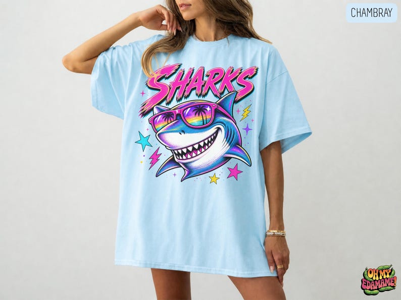 May include: A light blue t-shirt featuring a graphic of a shark wearing rainbow-tinted sunglasses with palm tree reflections. The word SHARKS is printed in large pink letters above the shark, surrounded by small colorful stars and lightning bolts.