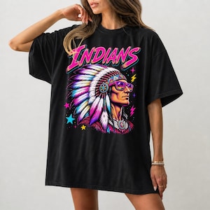 May include: A black t-shirt featuring a graphic print of a person wearing a large, colorful feathered headdress and purple-tinted sunglasses. The word INDIANS is printed in bright pink above the figure, surrounded by small blue and yellow stars.