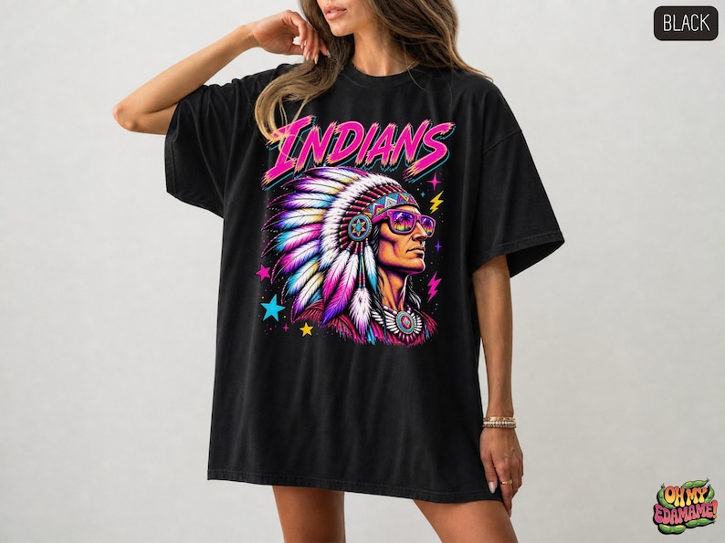 May include: A black t-shirt featuring a graphic print of a person wearing a large, colorful feathered headdress and purple-tinted sunglasses. The word INDIANS is printed in bright pink above the figure, surrounded by small blue and yellow stars.