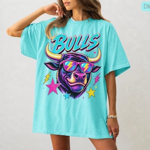May include: A chalky mint oversized t-shirt featuring a graphic of a bull wearing pink and yellow lightning bolt sunglasses. The word BULLS is printed in a stylized font above the bull, surrounded by colorful pink, yellow, and blue stars.