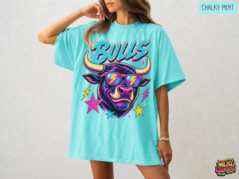 May include: A chalky mint oversized t-shirt featuring a graphic of a bull wearing pink and yellow lightning bolt sunglasses. The word BULLS is printed in a stylized font above the bull, surrounded by colorful pink, yellow, and blue stars.