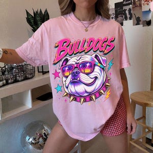 May include: A light pink t-shirt featuring a graphic of a bulldog wearing sunglasses with a palm tree print and a spiked collar. The text BULLDOGS is printed in a bold, stylized font above the dog, accompanied by colorful star and lightning bolt icons.