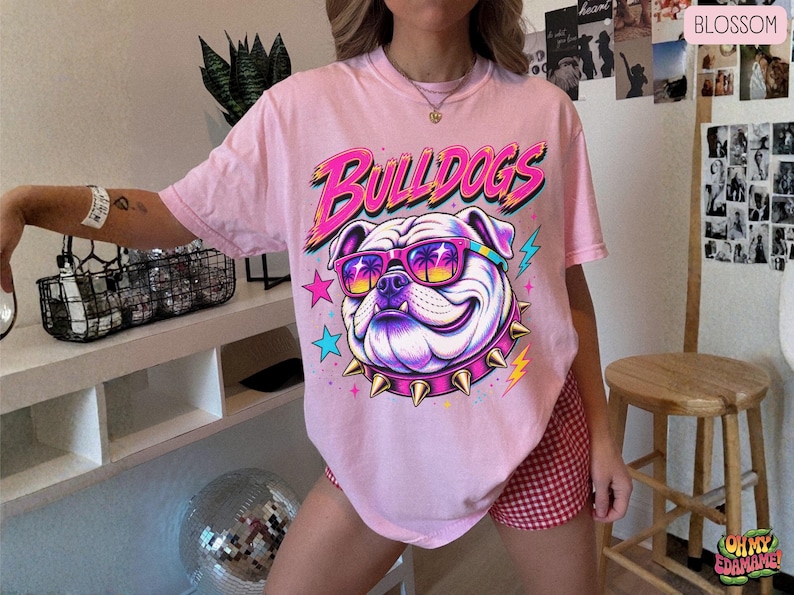 May include: A light pink t-shirt featuring a graphic of a bulldog wearing sunglasses with a palm tree print and a spiked collar. The text BULLDOGS is printed in a bold, stylized font above the dog, accompanied by colorful star and lightning bolt icons.