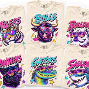 May include: Six ivory t-shirts featuring vibrant graphic prints of a tiger, bull, bulldog, panther, alligator, and shark. Each animal wears colorful sunglasses with palm tree reflections. The text Tigers, Bulls, Bulldogs, Panthers, Gators, and Sharks appears above each animal in a bold, stylized font with neon pink and blue accents.