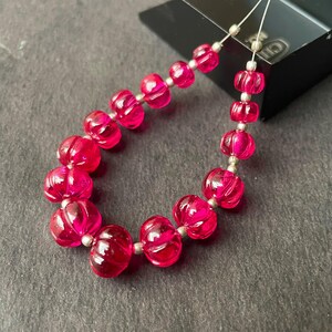 Puede incluir: Un hilo de cuentas translúcidas de color rosa intenso, acanaladas y con forma de calabaza, separadas por pequeñas cuentas espaciadoras de color plateado metálico. Las cuentas están dispuestas en forma de U, con las más grandes abajo.