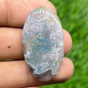 Puede incluir: Una gema ovalada con un patrón único de tonos blancos, grises, azules y rojos. La piedra tiene una superficie pulida y un aspecto natural. La gema se sostiene en la mano de una persona.