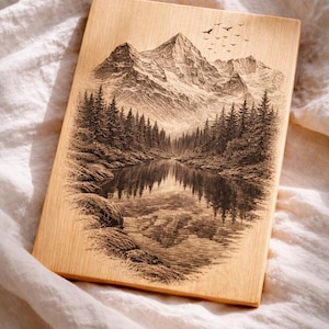 Puede incluir: Placa rectangular de madera con un grabado detallado de un paisaje montañoso con un lago y un bosque. La obra de arte está en tonos marrones y negros, con pájaros en el cielo. La placa descansa sobre una superficie blanca y texturizada. Medidas en centímetros.