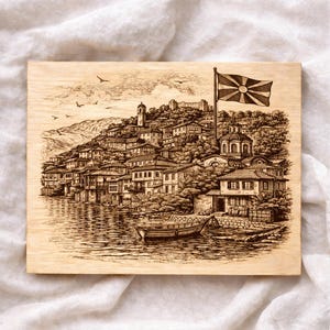 May include: A wood-burned art piece depicting a lakeside town with buildings, a church, and a flag. The artwork is in a detailed, vintage style, with a focus on the architecture and landscape. The flag is waving in the wind, adding a sense of movement to the scene.