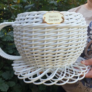Könnte beinhalten: Ein weißer, geflochtener Weidenkorb in Form einer großen Teetasse mit passendem Untertassenboden und einem geschwungenen Henkel. Auf dem vorderen Rand befindet sich ein kleines goldenes Schild mit der Aufschrift Rattan Garden in schwarzer Schrift.
