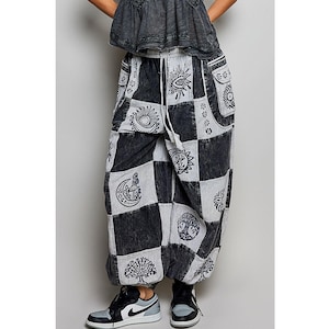May include: Black and white patchwork pants with a drawstring waist. The pants feature various celestial and nature-inspired designs, including suns, moons, and trees. The pants are paired with black and gray sneakers.