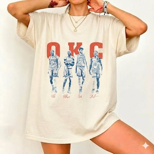 May include: Cream-colored t-shirt featuring the text "OKC" in red block letters above a blue and white illustration of four basketball players. The players' signatures are below their images. The shirt has short sleeves and a relaxed fit.