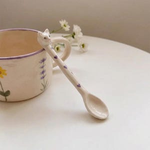 May include: A ceramic spoon with a cat head handle and purple heart accents. The spoon is next to a floral decorated mug with a purple rim. The mug has a yellow flower and lavender sprig design.